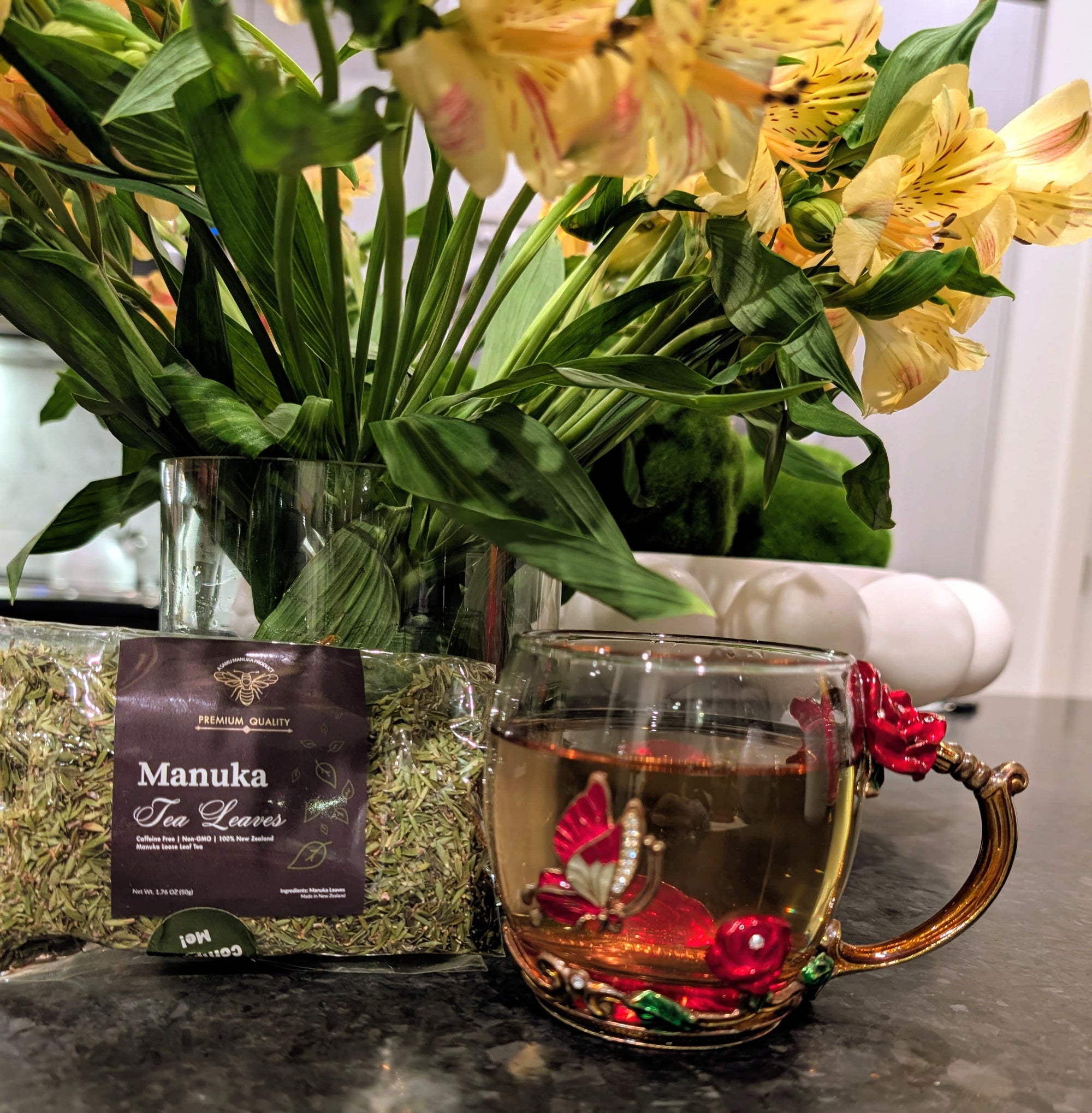 Tea cup with floral design, Manuka Tea Leaves packaging, and flowers on a dark surface.